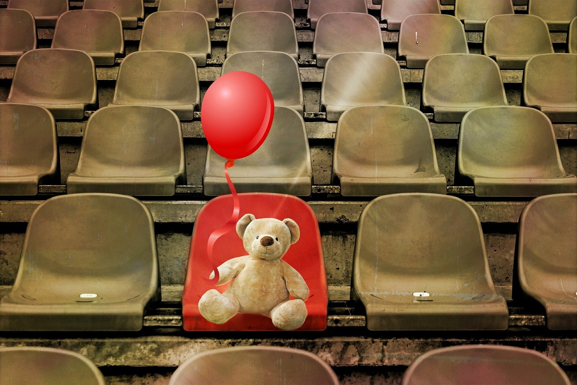 sedie di una tribuna vuote e marroni, solo una rossa occupata da un orsacchiotto che tiene in mano un palloncino rosso