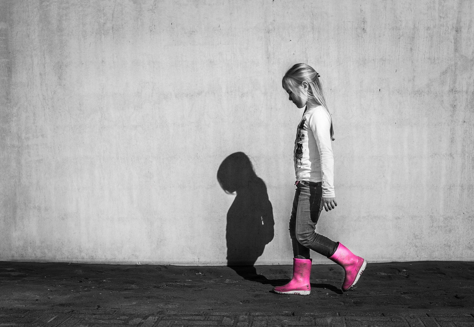 foto in bianco e nero, una bimba cammina a testa bassa accanto ad un muro che riflette la sua ombra, con stivaletti rosa